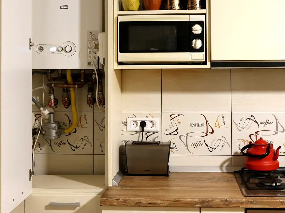 Caldera mural de gas dentro de un mueble de cocina abierto, con sus tuberías y llaves debajo, junto a un microondas y una placa de gas
