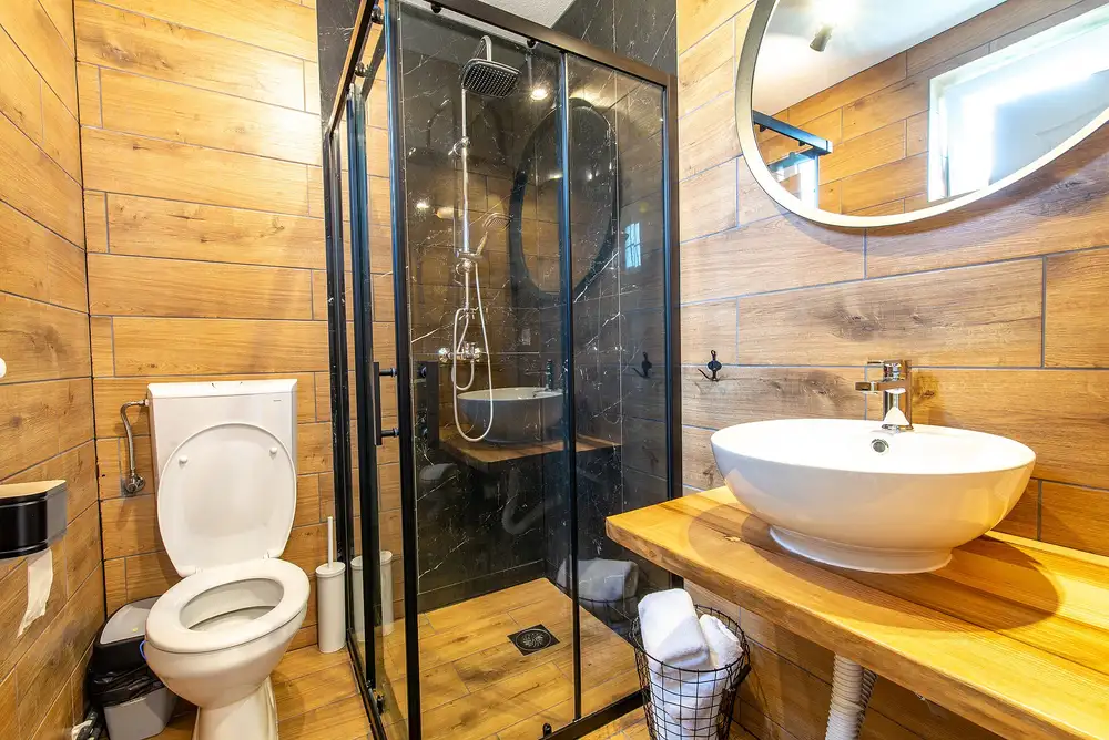 Baño con ducha de cristal de perfil negro, paredes de imitación madera, lavabo sobre encimera y espejo redondo