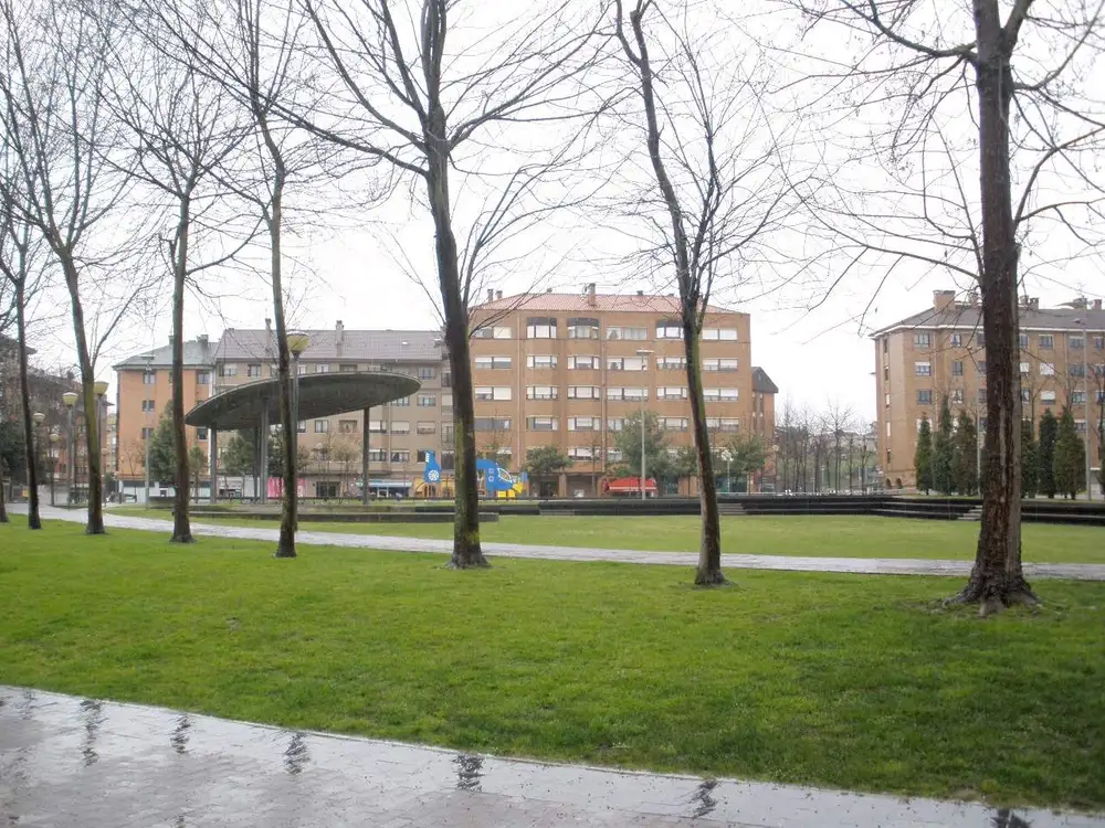 Parque con césped y árboles sin hojas en Lugones, con una pérgola circular y bloques de pisos de ladrillo al fondo en un día de lluvia