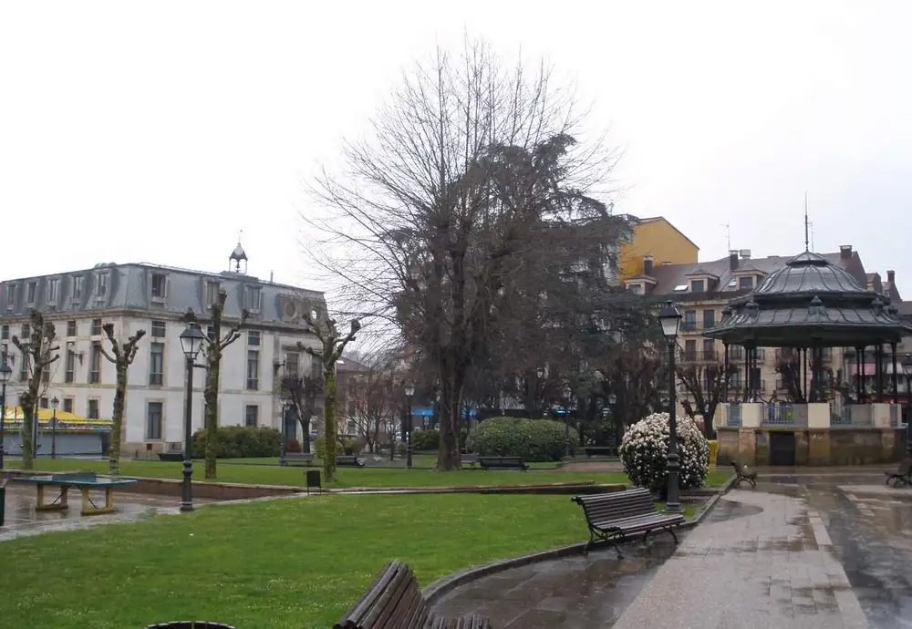Parque con césped, bancos y un gran árbol en la Pola de Siero, con un templete de música y edificios de varias plantas al fondo en un día de lluvia